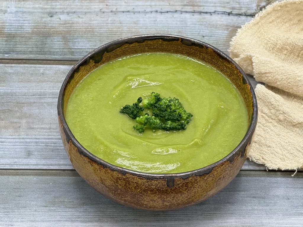 A bowl of creamy broccoli soup garnished with steamed broccoli, served in a brown ceramic bowl on a rustic wooden table.